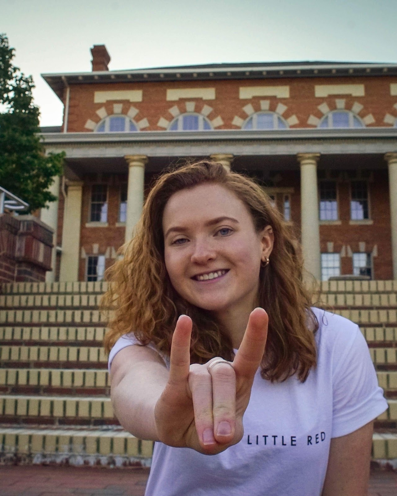 Lauren White makes a Wolfie sign with her hand in front of a building on campus