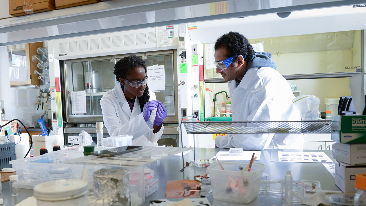 Two students in white lab coats examine a sample in a lab.