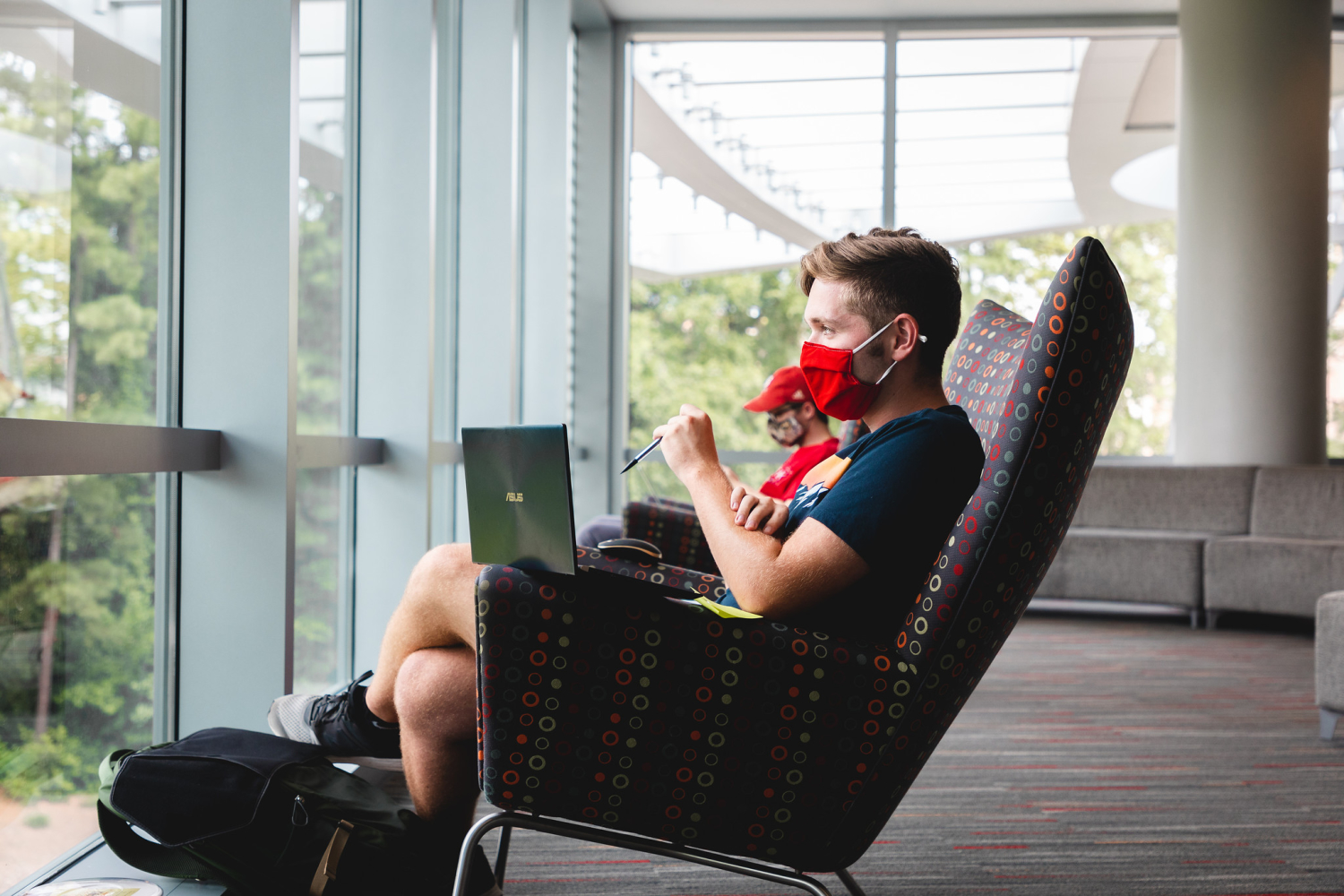 A masked, male student with a laptop in his lap looks out the window in Talley Student Union