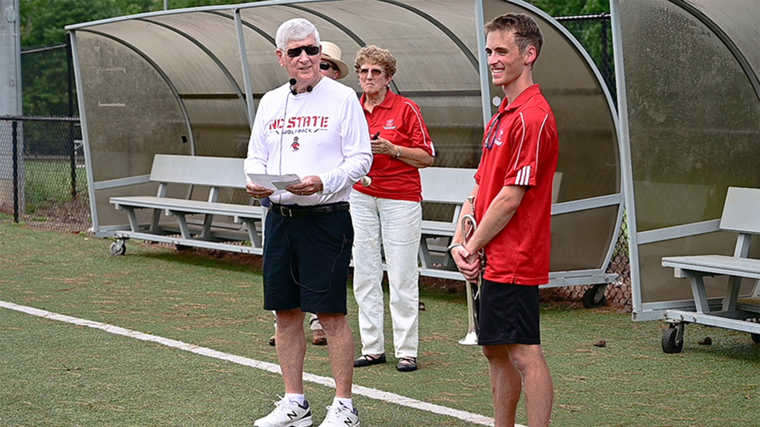 Gage Fringer, wearing a red shirt and black shorts and holding his trumpet, standing next to a man (Bob Masini) in a white shirt and black shorts