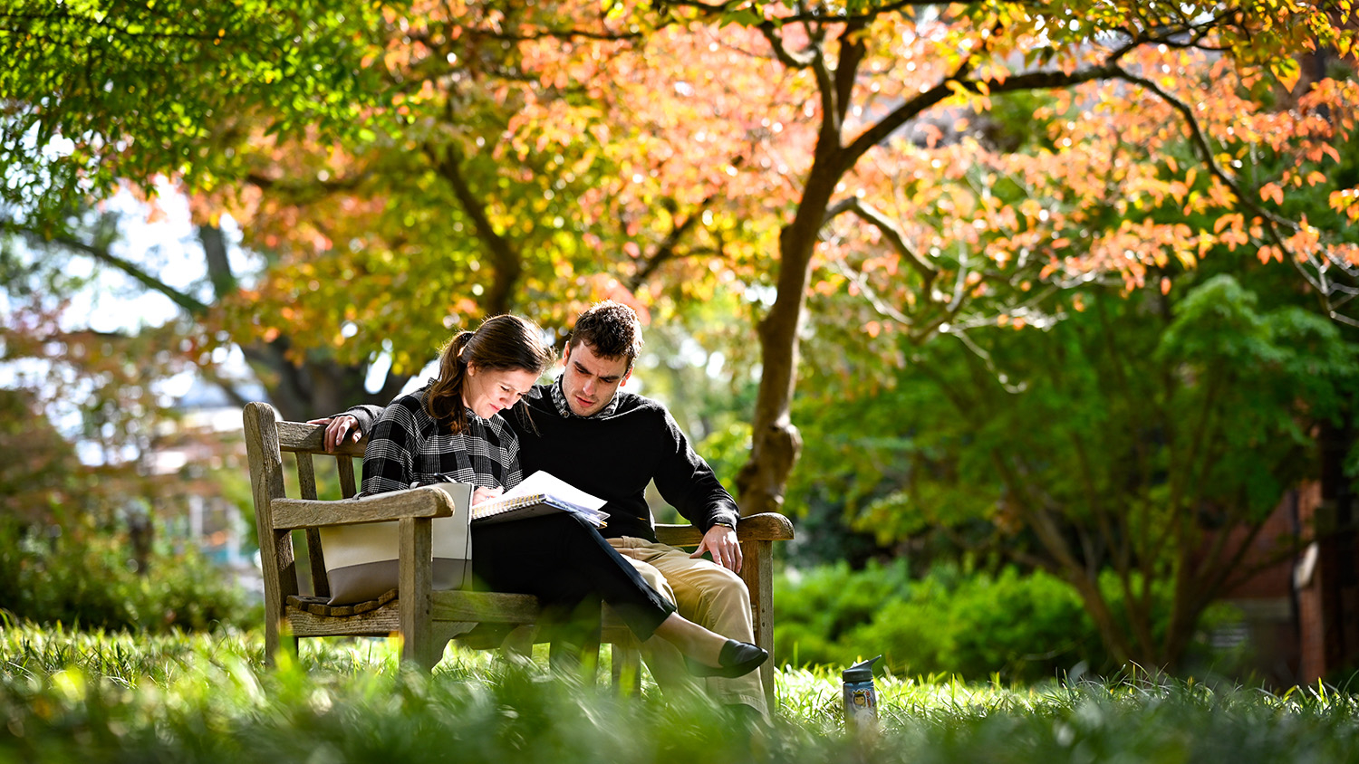 Two people sitting by side on an outdoor bench, looking over a notebook.
