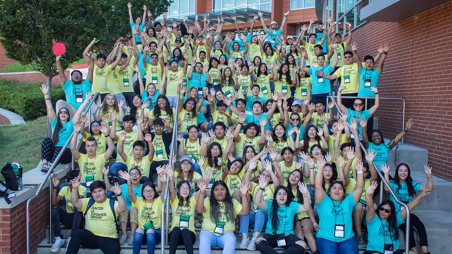 A large group of students in teal and yellow shirts raising their hands