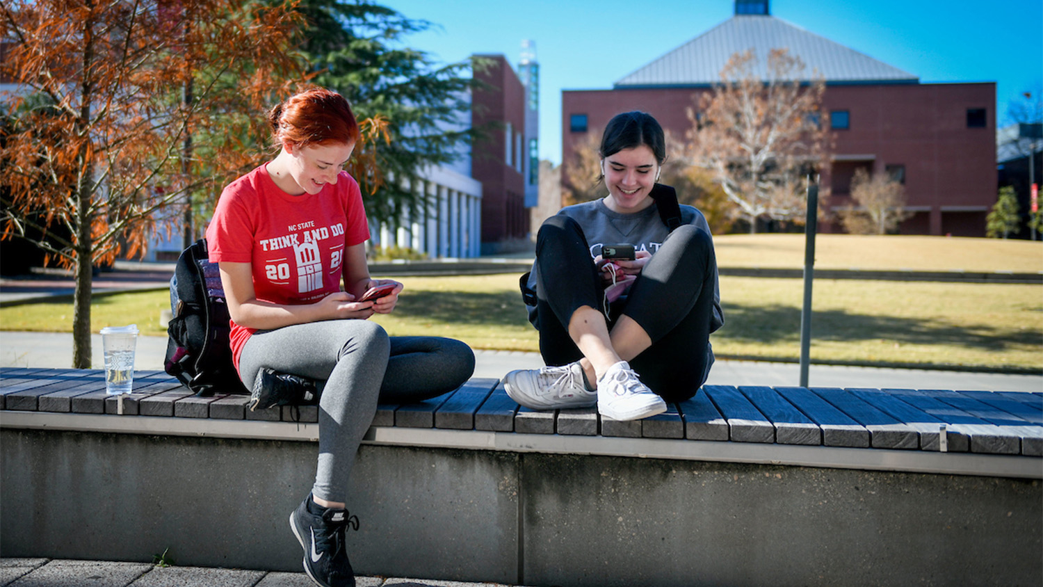 Two students smiling and sitting on a concrete wall while looking at their phones