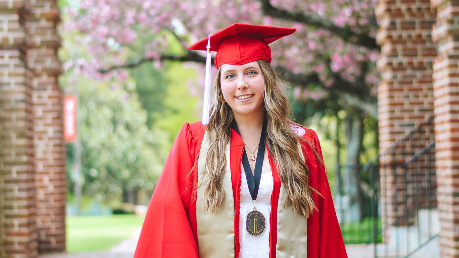 Lindsay Strickland in red graduation robes and hat in front of the brick columns at the front of Holladay Hall