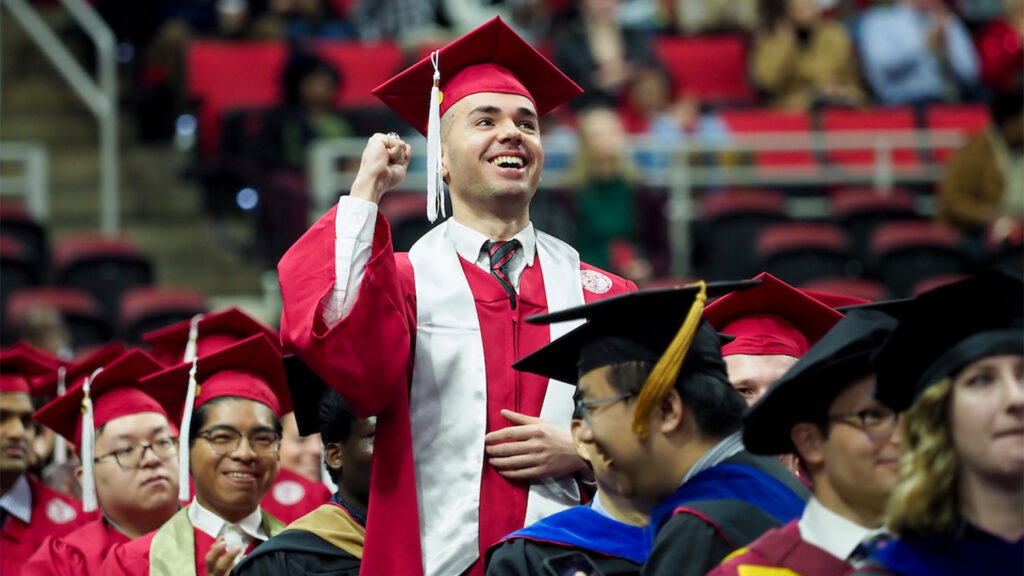 A male graduate in red robes stands up in a crowd of seated graduates and makes a fist pump sign