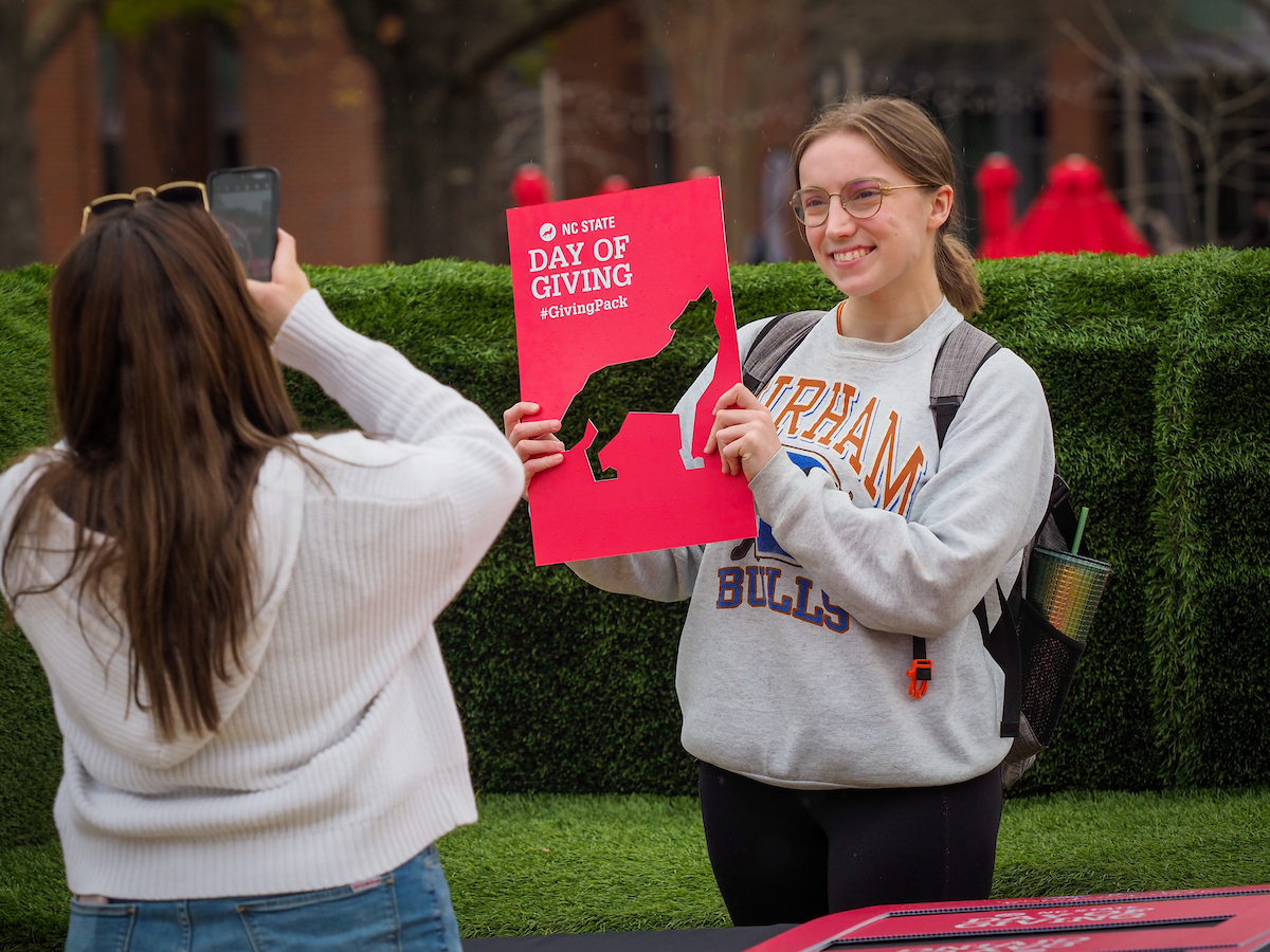 A female student takes another student's picture while she holds a sign that reads "Day of Giving."