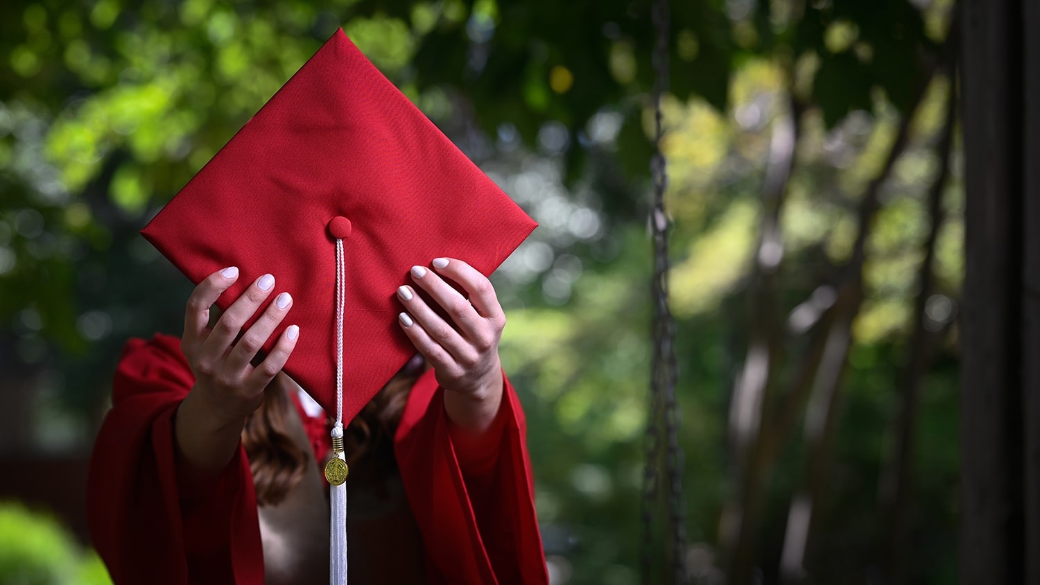 A student wearing a red graduation gown holds up her mortar board in front of her face.