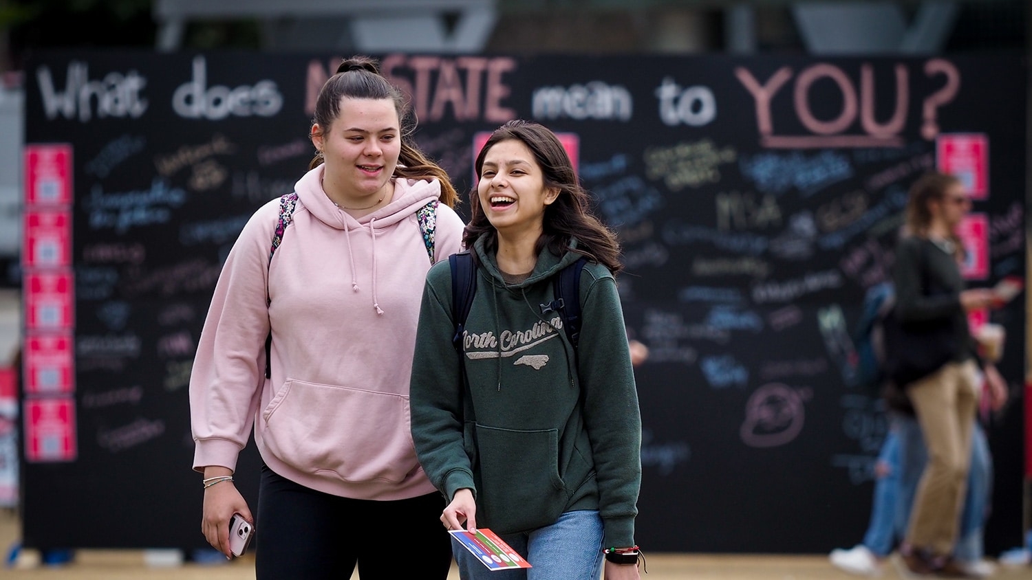 Two students laugh together while walking in front of a chalkboard sign that reads "What does NC State mean to you?" with hand written comments below the question.