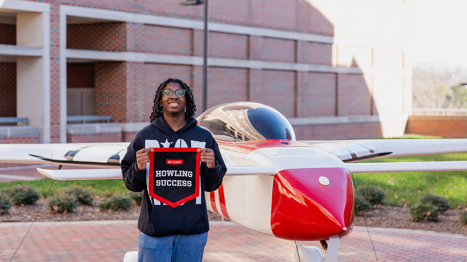 Caston Reaves poses with his Howling Success banner in front of a model plane outside Engineering Building III on Centennial Campus.