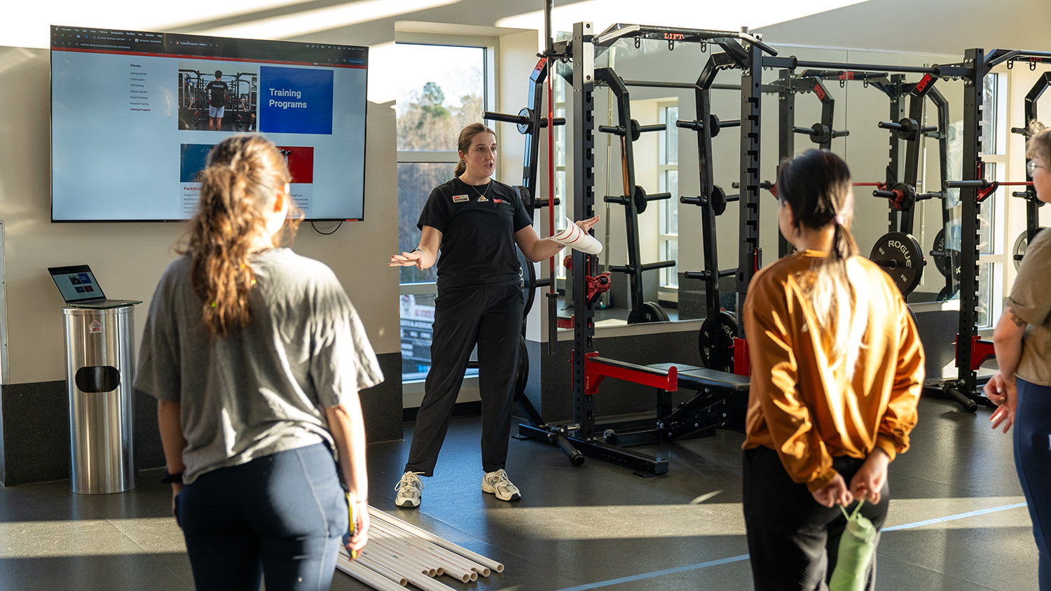 A Wellness and Recreation staff member speaks to students in front of several weight machines.