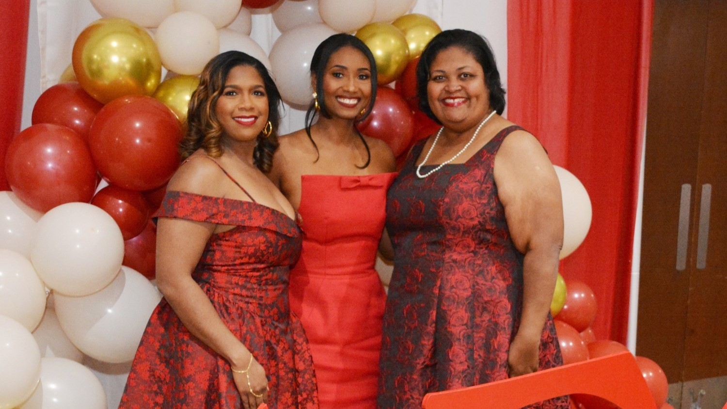 Three women in red dresses posing in front of balloons
