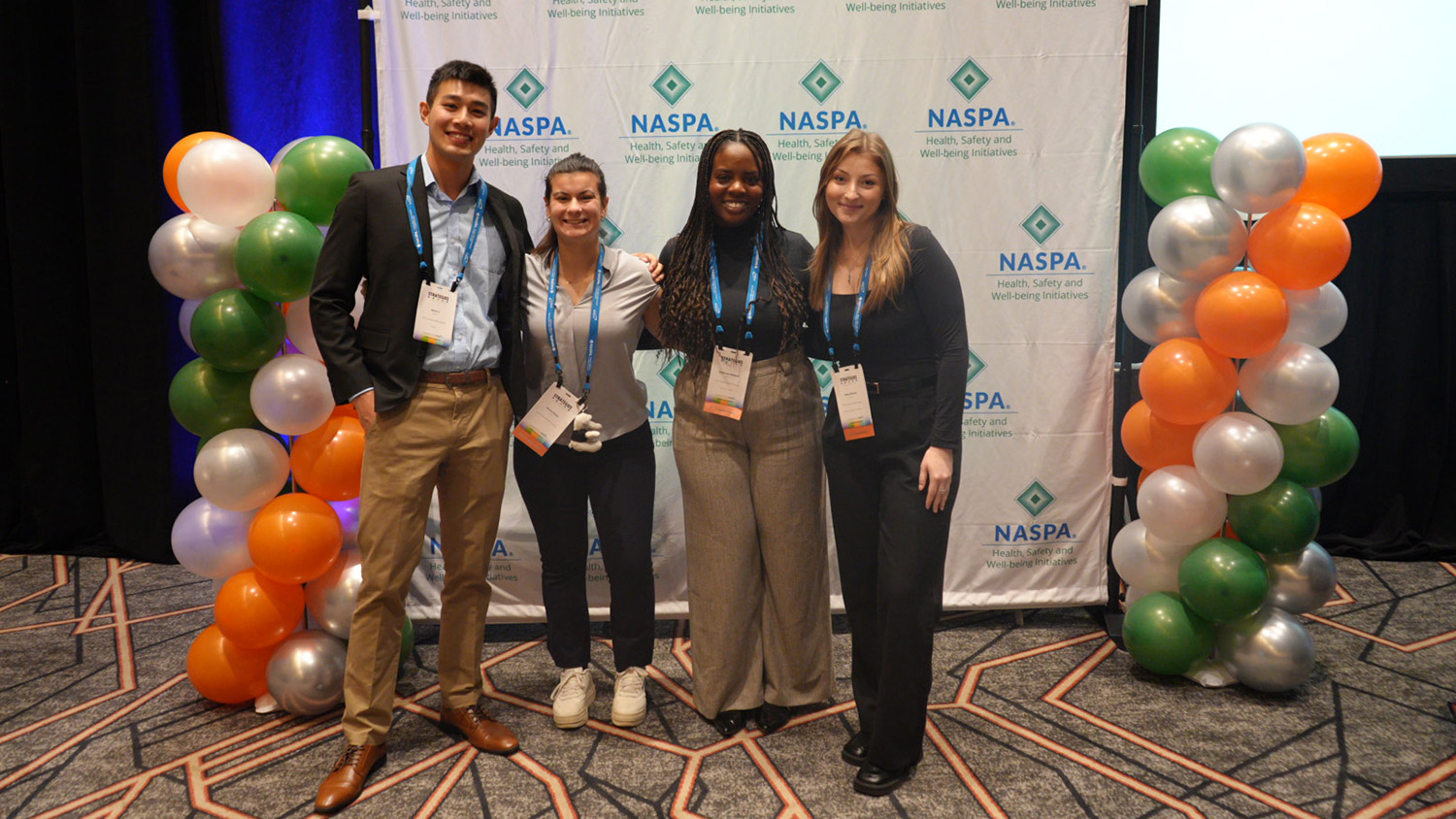 From left: Pack Peers students Devin Li, Emma Despa, Ore Badejoh and Haley Berland in front of a NASPA banner and two white, green and silver balloon structures at the NASPA conference in Chicago.